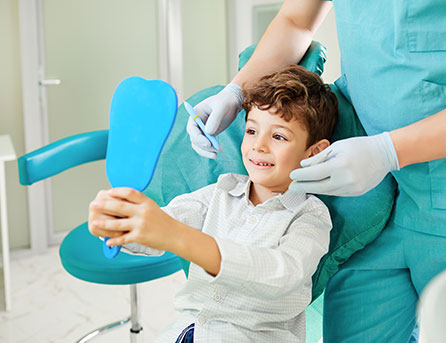 Child smiling while holding a mirror in a dental chair, with a dentist in gloves preparing for a fluoride treatment, emphasizing pediatric dental care at Citrus Smiles Dental Studio.