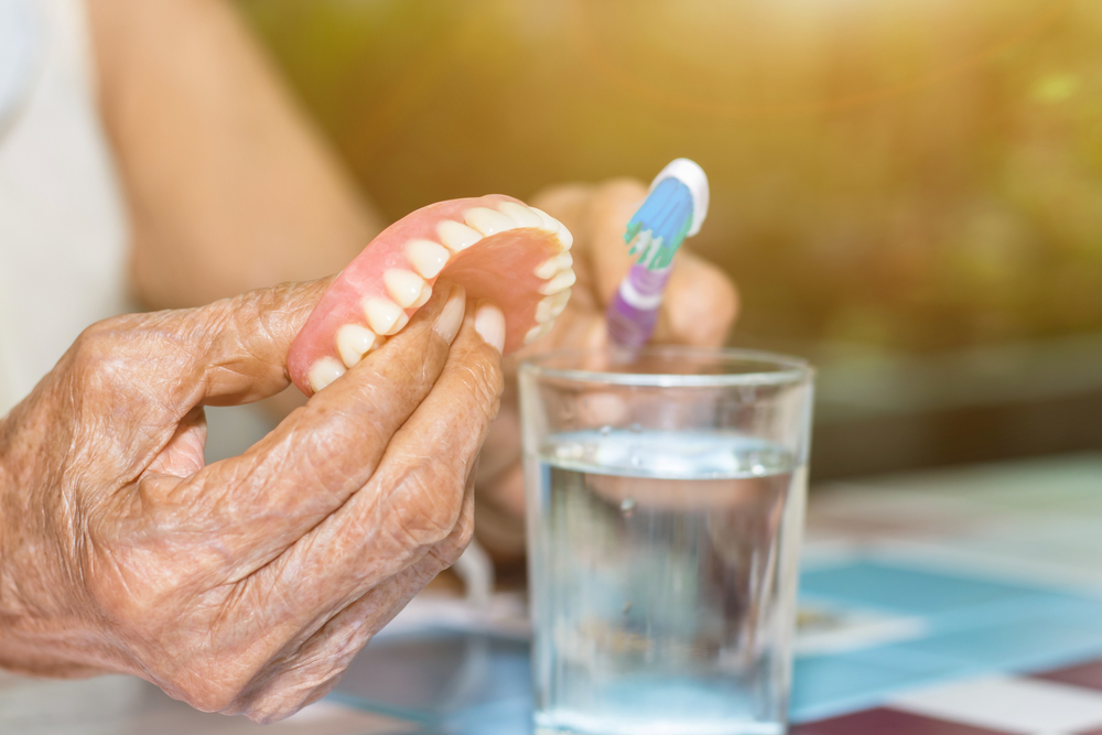 Old,Women,Hand,Hold,Fake,Teeth,With,Dirty,For,Removable Elderly person holding dentures and toothbrush near a glass of water, emphasizing denture care and maintenance for oral health.