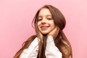 Girl smiling confidently with metal braces against a pink background, showcasing orthodontic treatment benefits for teens.